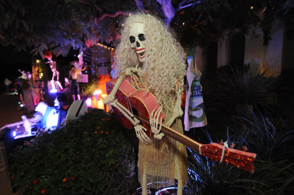 A skeleton wearing a dress and playing a guitar outside of a home.
