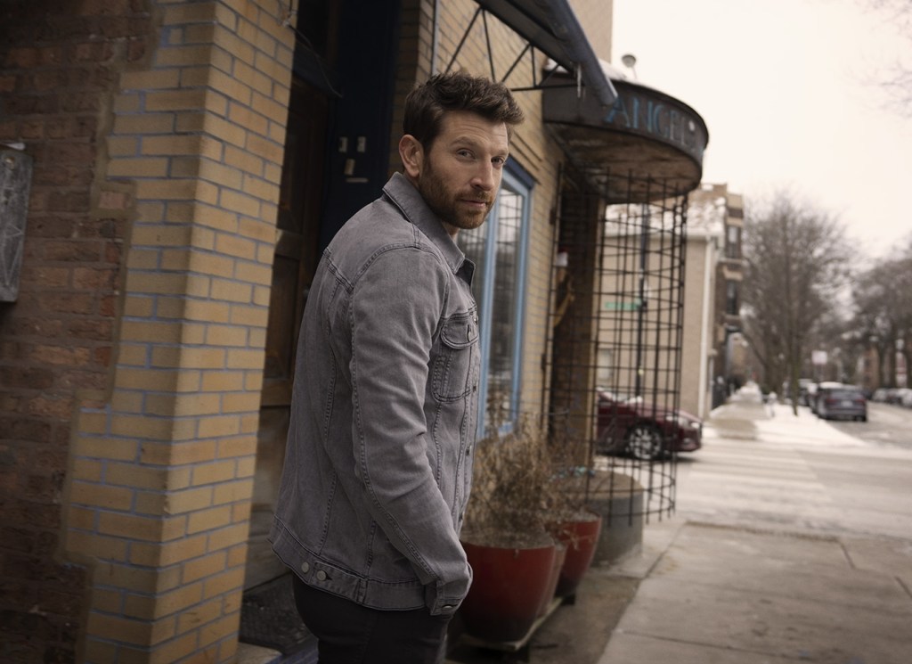 A bearded man in a jeans jacket looks over his shoulder on a street.
