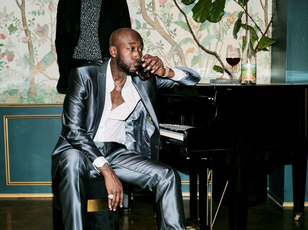 A bald Aftrican-American man sips a drink while sitting at a piano.
