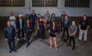 A group of 13 musicians standing in front of a gray wall.