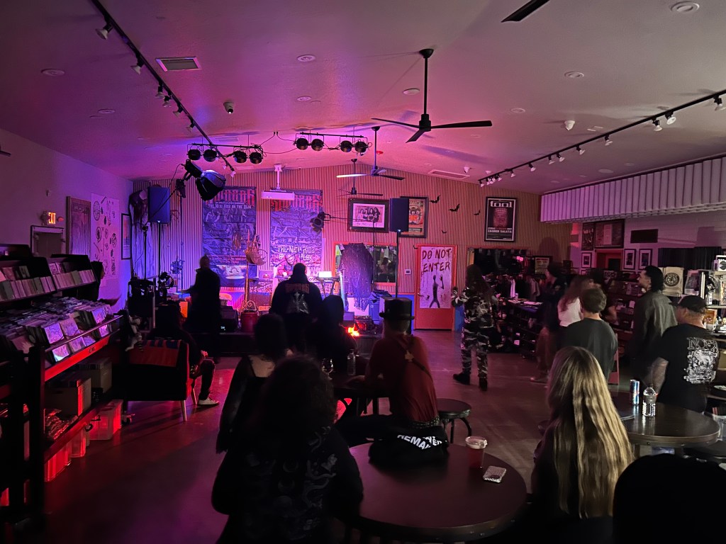 A crowd watches as a band performs onstage in a room with purple lighting.