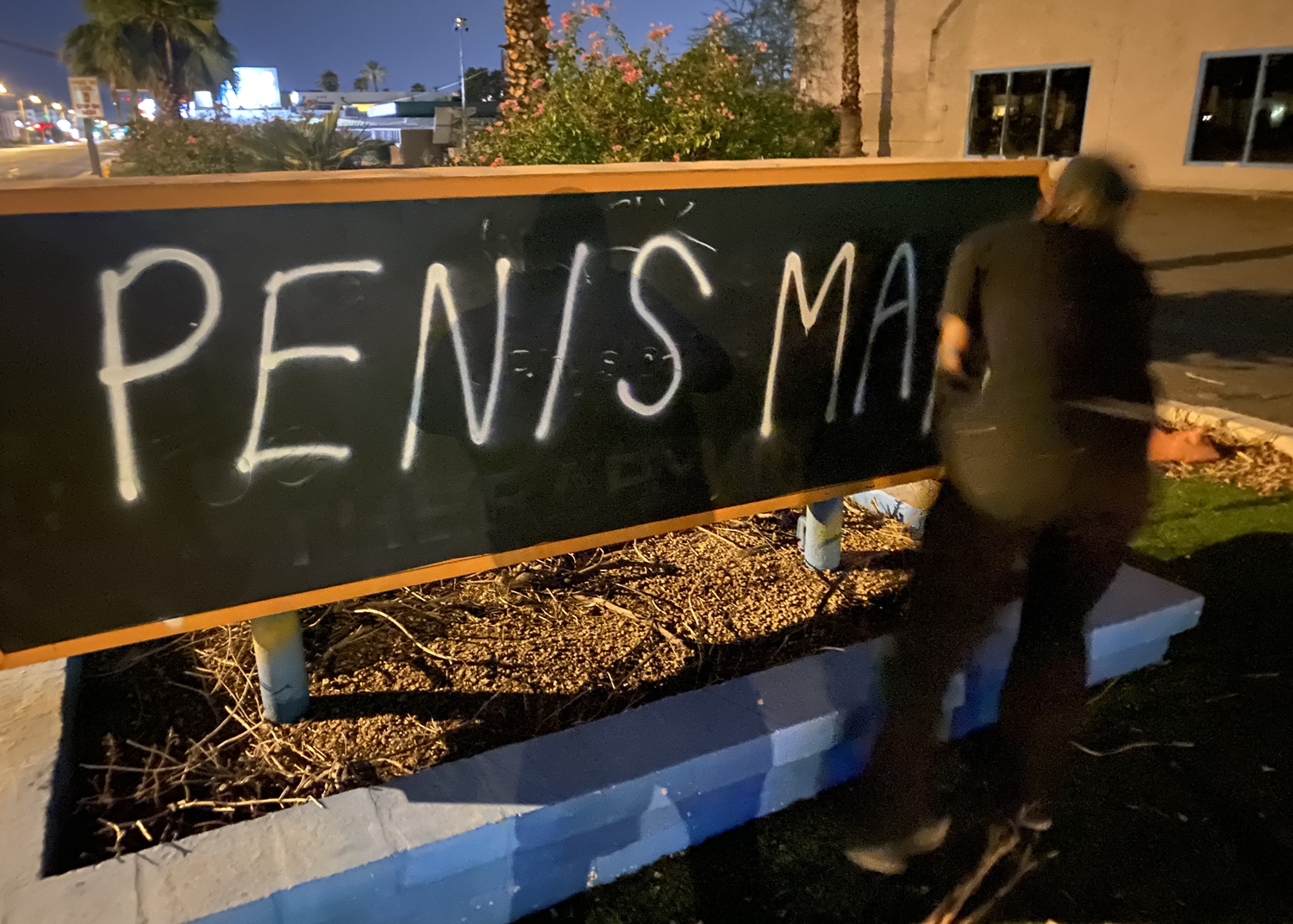 A male dressed in black clothing spray paints the words "PENIS MAN" on a sign.