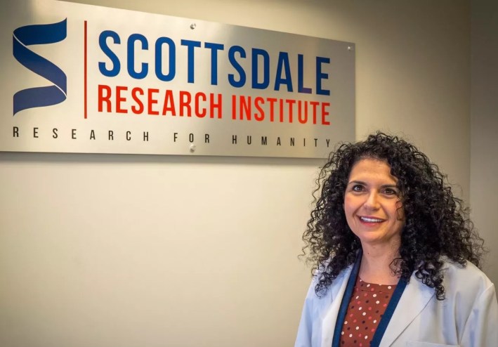 a woman in a lab coat stands in front of a sign for Scottsdale Research Institute