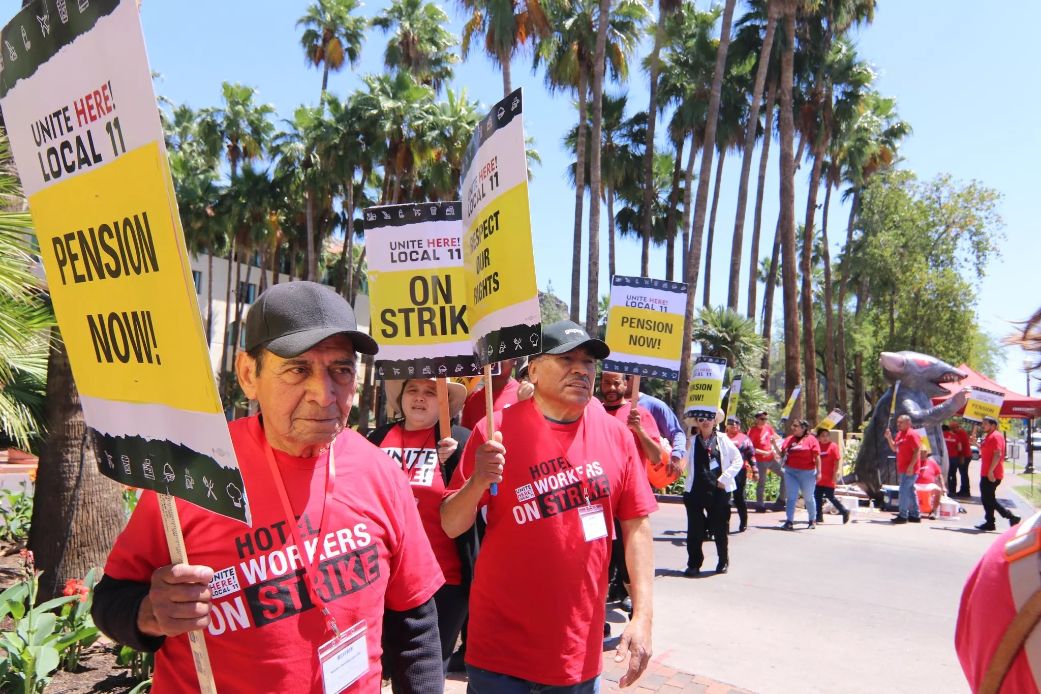 Hotel workers in red shirts hold picket signs with phrases like "On Strike!" and "Pension Now!" In the background is an inflatable rat.