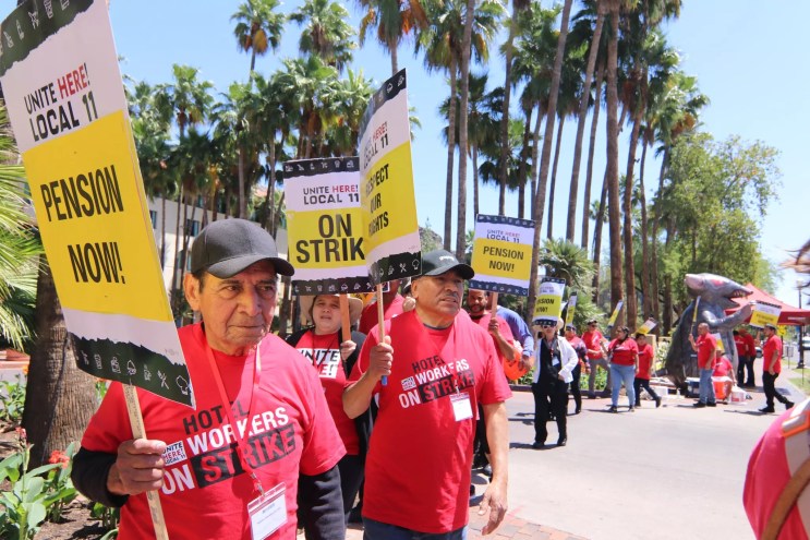Hotel workers in red shirts hold picket signs with phrases like "On Strike!" and "Pension Now!" In the background is an inflatable rat.