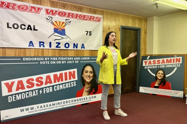 Yassamin Ansari, in a yellow blazer, speaks in front of campaign banners with her name and face on them.