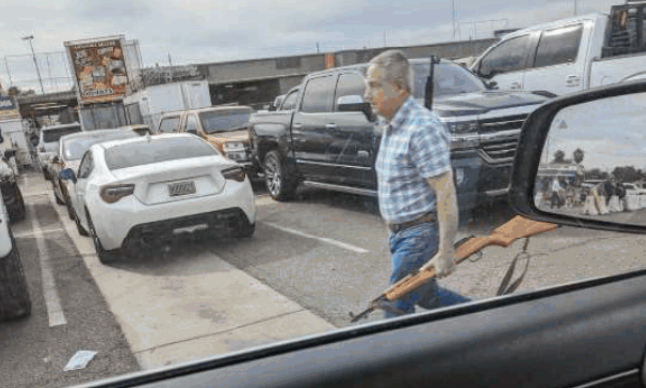 A man holds a rifle as he walks by a car, as seen from within the vehicle