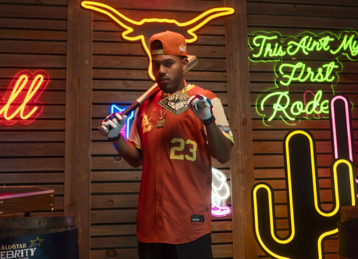 A man in a baseball jersey in front of a wall of neon signs.
