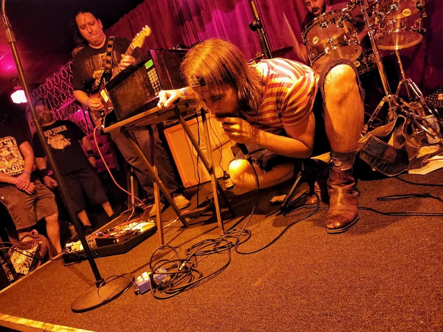 A long-haired man crouches on a stage while holding and singing into a microphone.