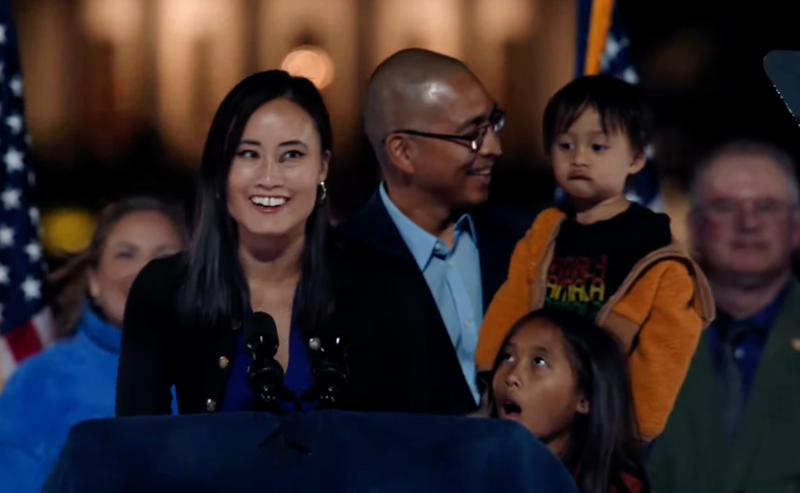 a woman speaks at a podium with her husband and two children behind her