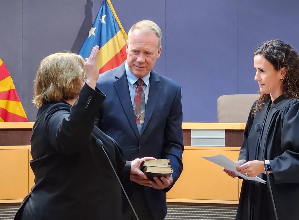 A woman raising her right hand with her left on a bible in front of a female judge. A gray-haired man holds the bible