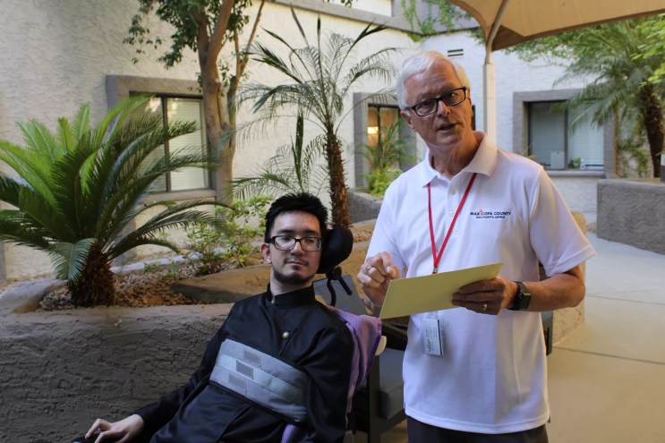 a young man in a wheelchair listens as an older man in a while shirt talks