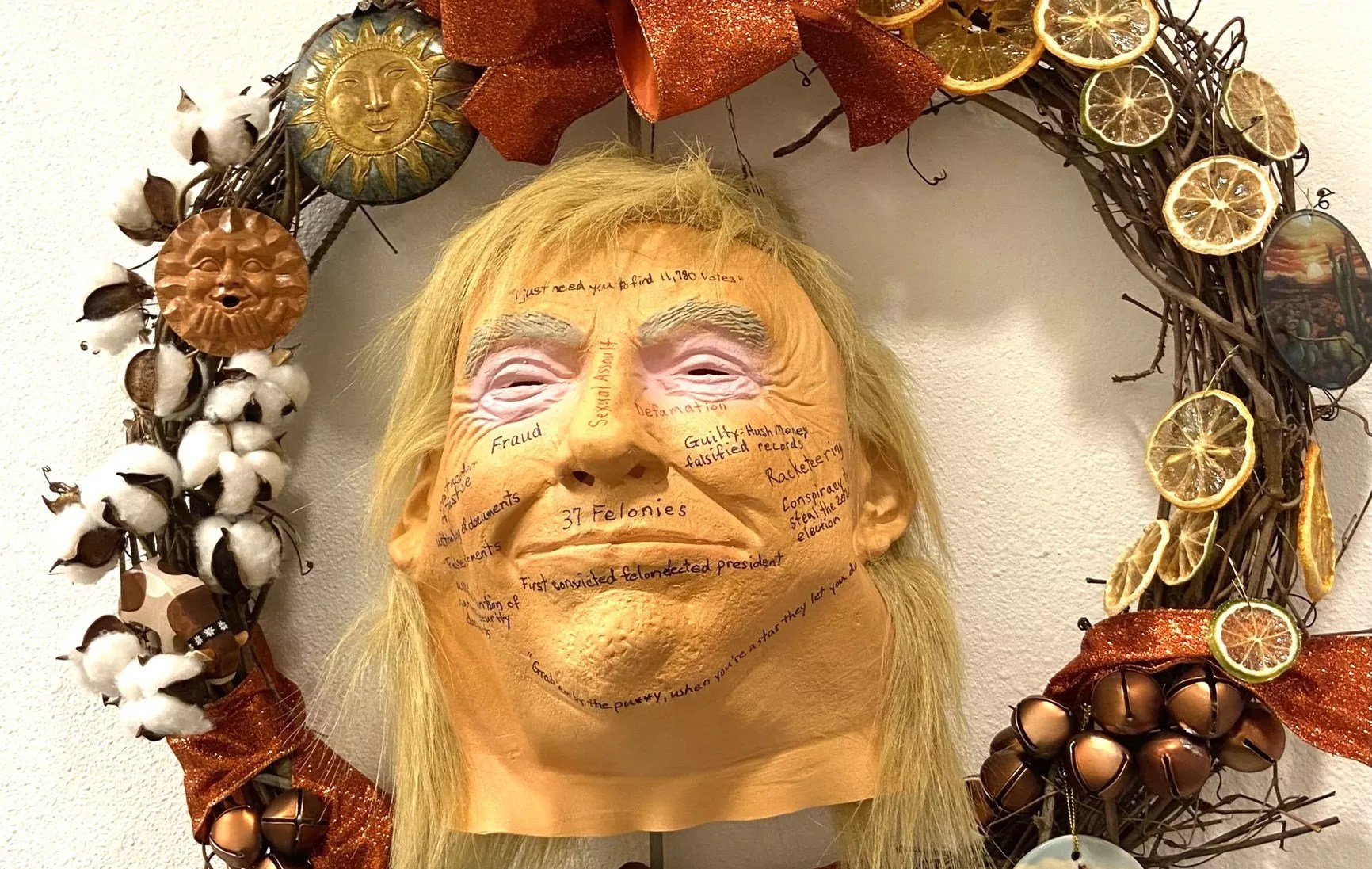a wreath featuring a rubber trump mask with phrases like "defamation" and "sexual assault" written on its face