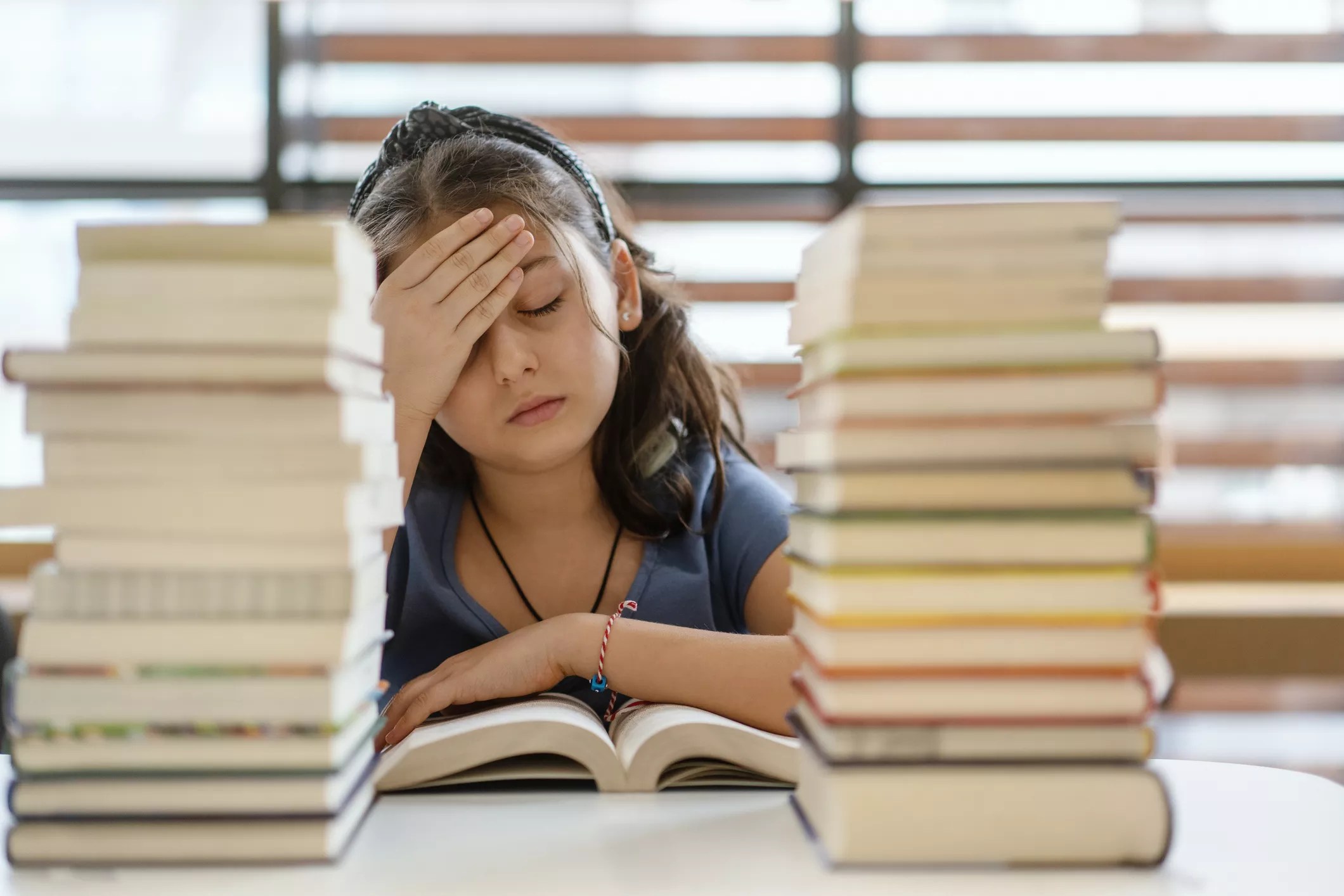 an elementary school girl puts a hand on her head in exasperation while stacks of books sit in front of her
