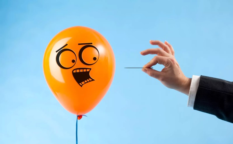 a balloon with a scared face looks at a hand with a pin