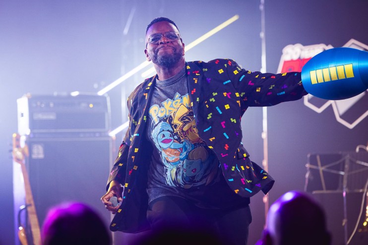 An African-American man in a Pac-Man themed jacket stands on stage with a toy cannon covering his left arm.