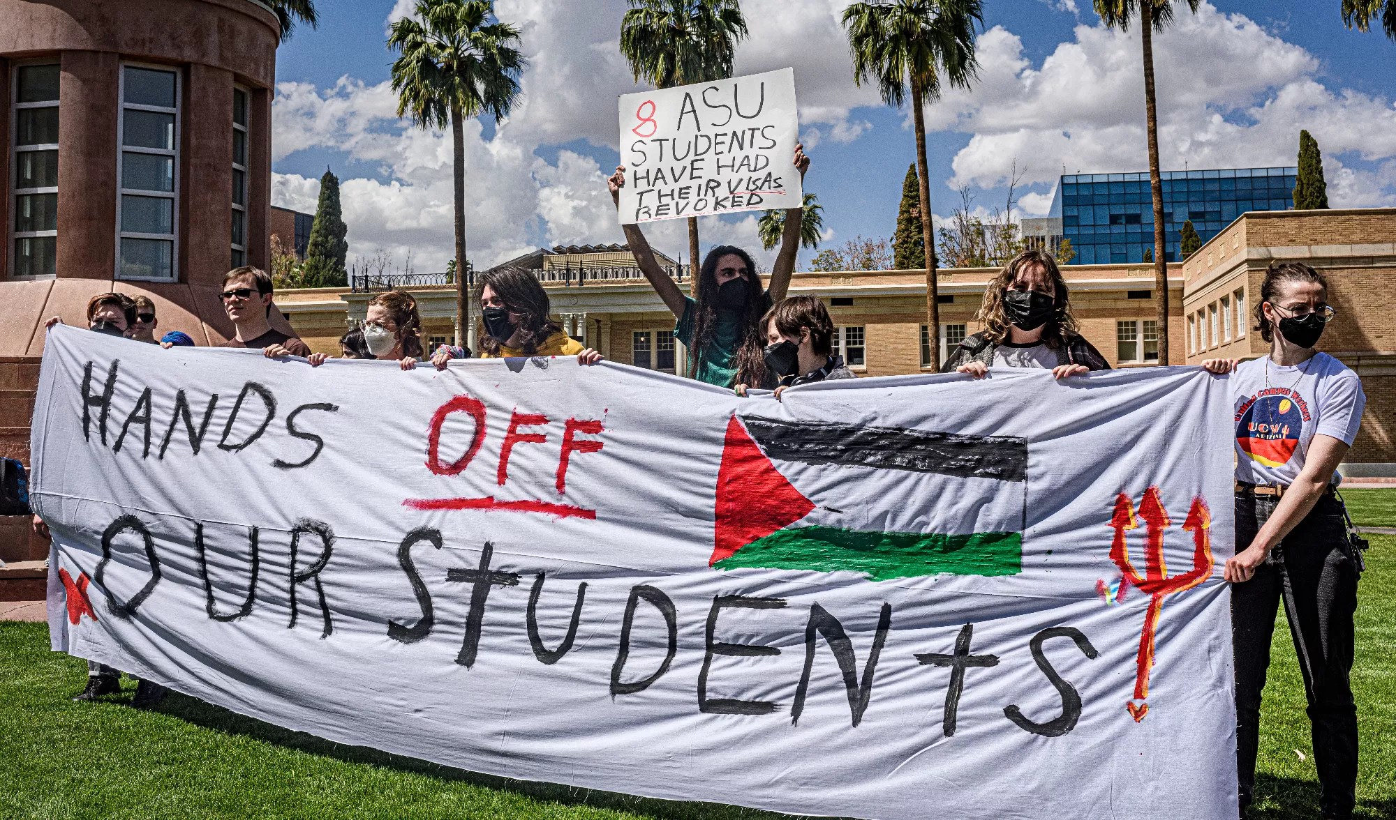 people hold a banner that says "hands off our students" with a Palestinian flag