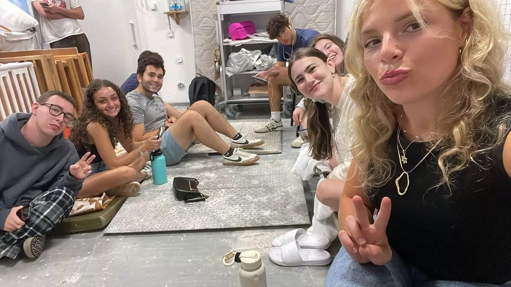 young adults sit on the floor and pose for a selfie in a bomb shelter