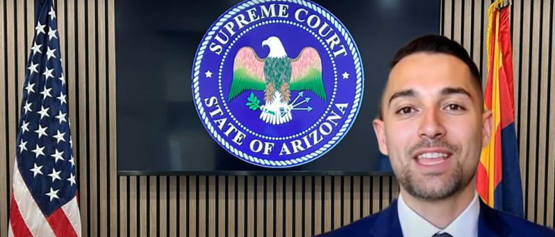 an ai-generated man in a suit speaks to the camera in a "room" with the U.S. and Arizona flags and a TV with the Arizona Supreme Court seal on it