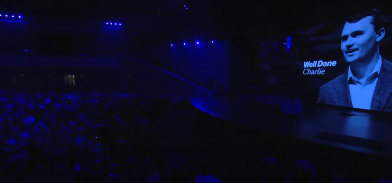 a darkened auditorium with a screen featuring charlie kirk and the words "well done charlie"